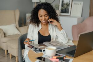 A woman multitasks with a laptop and magazine at a cozy home office, sipping coffee.