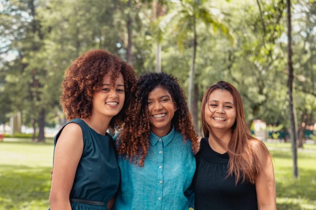Three women with diverse hairstyles smiling outdoors in a park setting.
